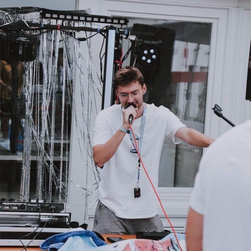 Janik Rabenstein strahlt vor Begeisterung, während er das Mikrofon in der Hand hält und die magische Atmosphäre der unvergesslichen Feldparty einfängt. Die lebendigen Lichter im Hintergrund unterstreichen die einzigartige Stimmung dieses mitreißenden Events.