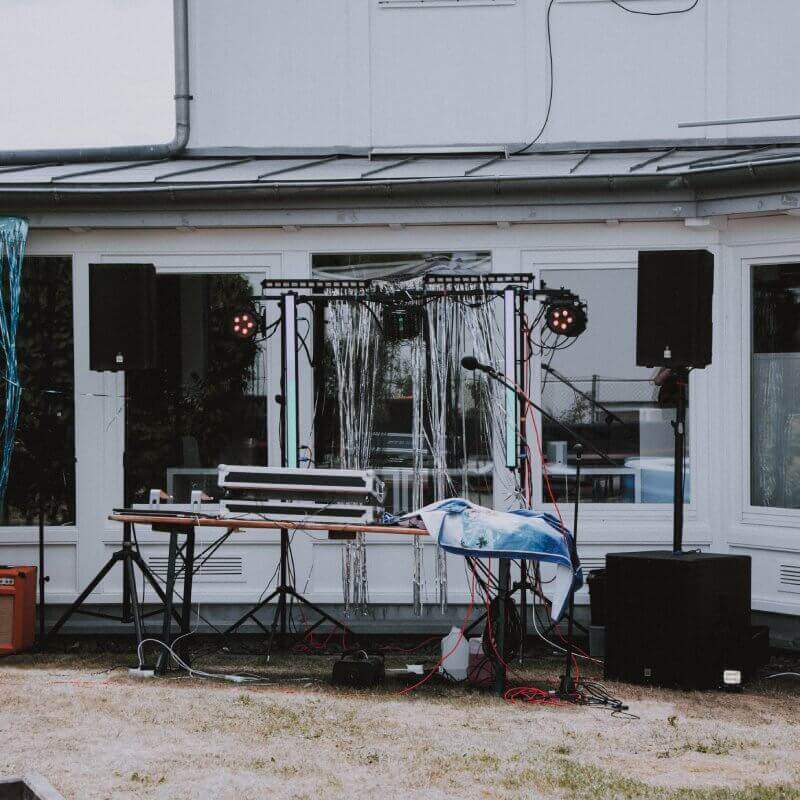 Die Sonne strahlt und taucht die Szenerie in warmes Licht, während die beeindruckenden Lautsprecher und funkelnden Lichter das Sommerfest mit energiegeladener Musik zum Leben erwecken. Vor dem imposanten Tisch mit dem DJ-Controller entsteht eine mitreißende Atmosphäre, die die Vorfreude auf eine unvergessliche Party steigert.